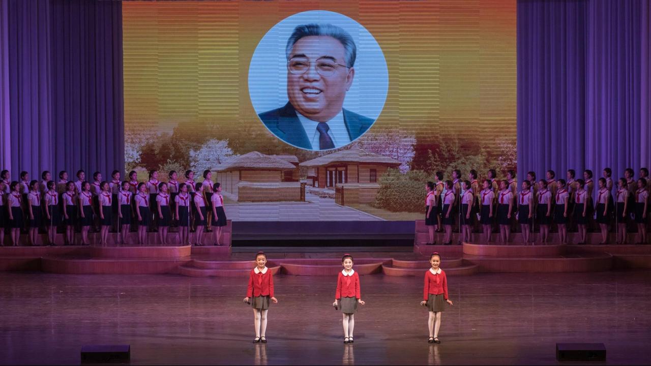 Mädchen singen und tanzen auf einer Bühne, im Hintergrund ein überdimensionales Porträt von Kim Il Sung.