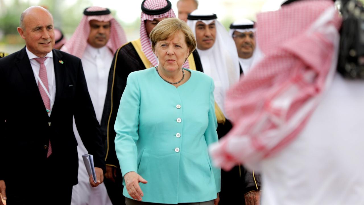 Bundeskanzlerin Merkel am Flughafen von Dschidda in Saudi-Arabien. Sie trägt einen türkisen Blazer und ist von Männern umgeben.