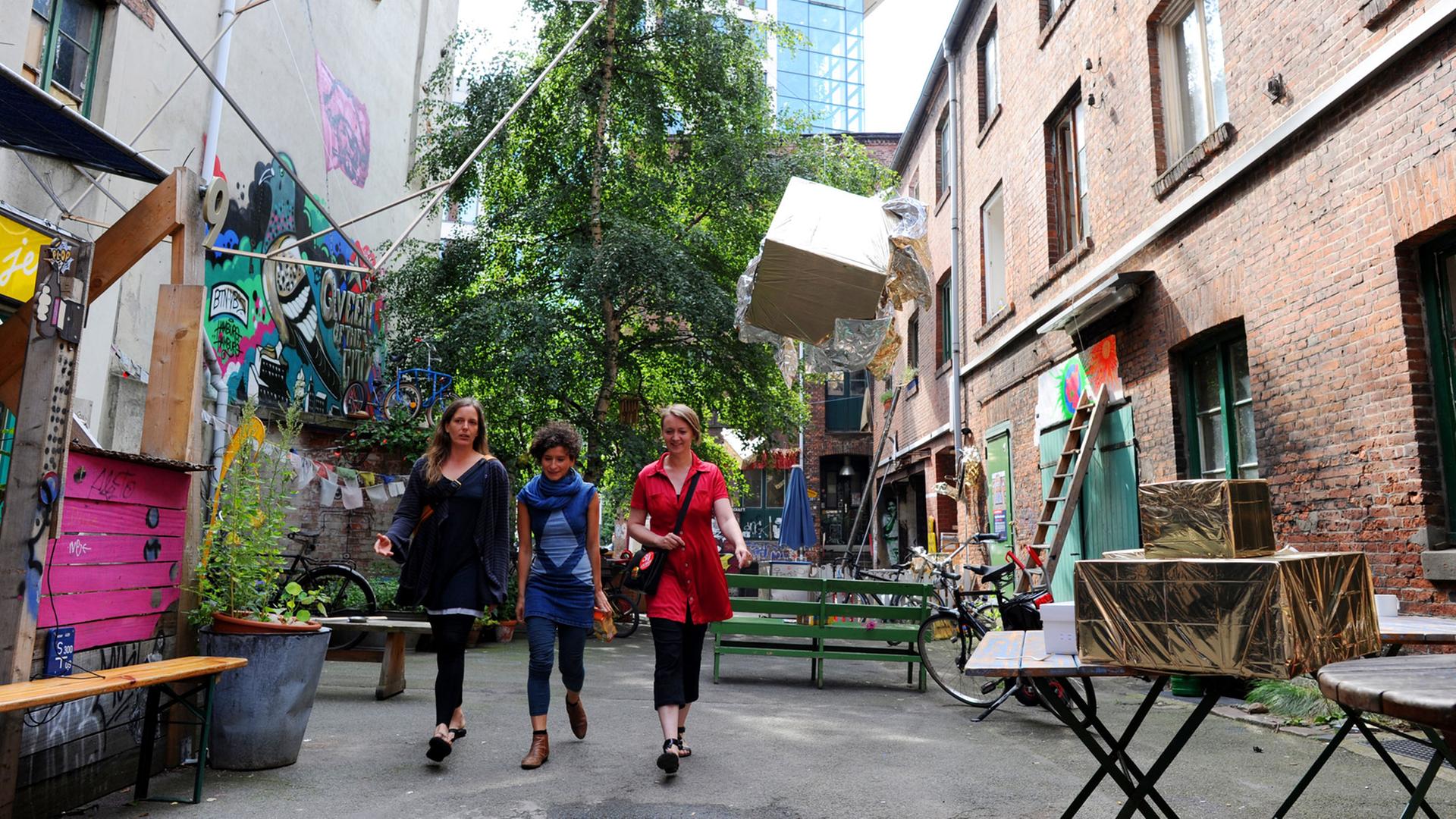 Drei Frauen laufen nebeneinander durch einen allerlei buten Gegenständen bestückten Hof, rundherum Altbauten.