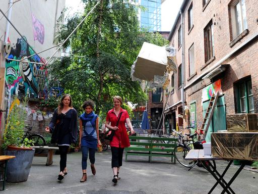 Drei Frauen laufen nebeneinander durch einen allerlei buten Gegenständen bestückten Hof, rundherum Altbauten.
