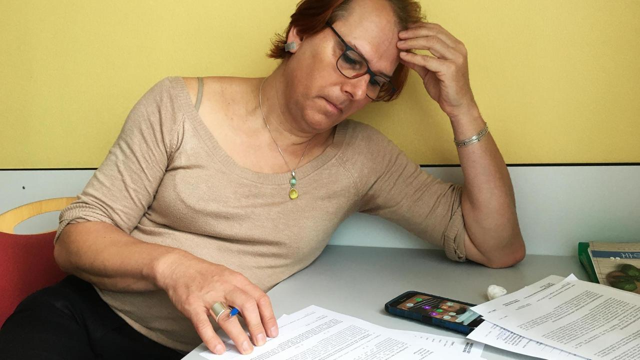 Elna sitzt an einem Krankenhaustischchen vor einer gelben Wand, Kopf auf die linke Hand aufgestützt, und liest Dokumente. Elna sitzt an einem Krankenhaustischchen vor einer gelben Wand, Kopf auf die linke Hand aufgestützt, und liest Dokumente.