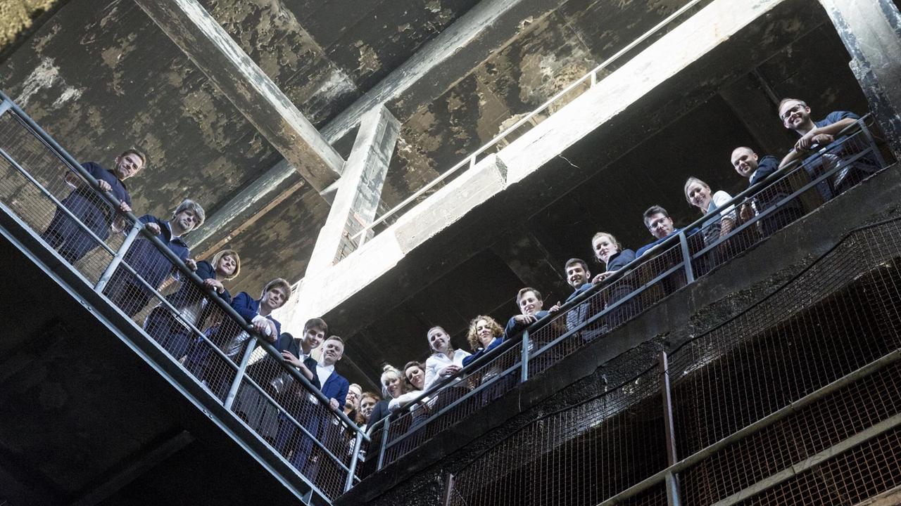 Der Chor schaut von einer Brüstung in einem alten Werk runter in die Kamera. Der Chor schaut von einer Brüstung in einem alten Werk runter in die Kamera.