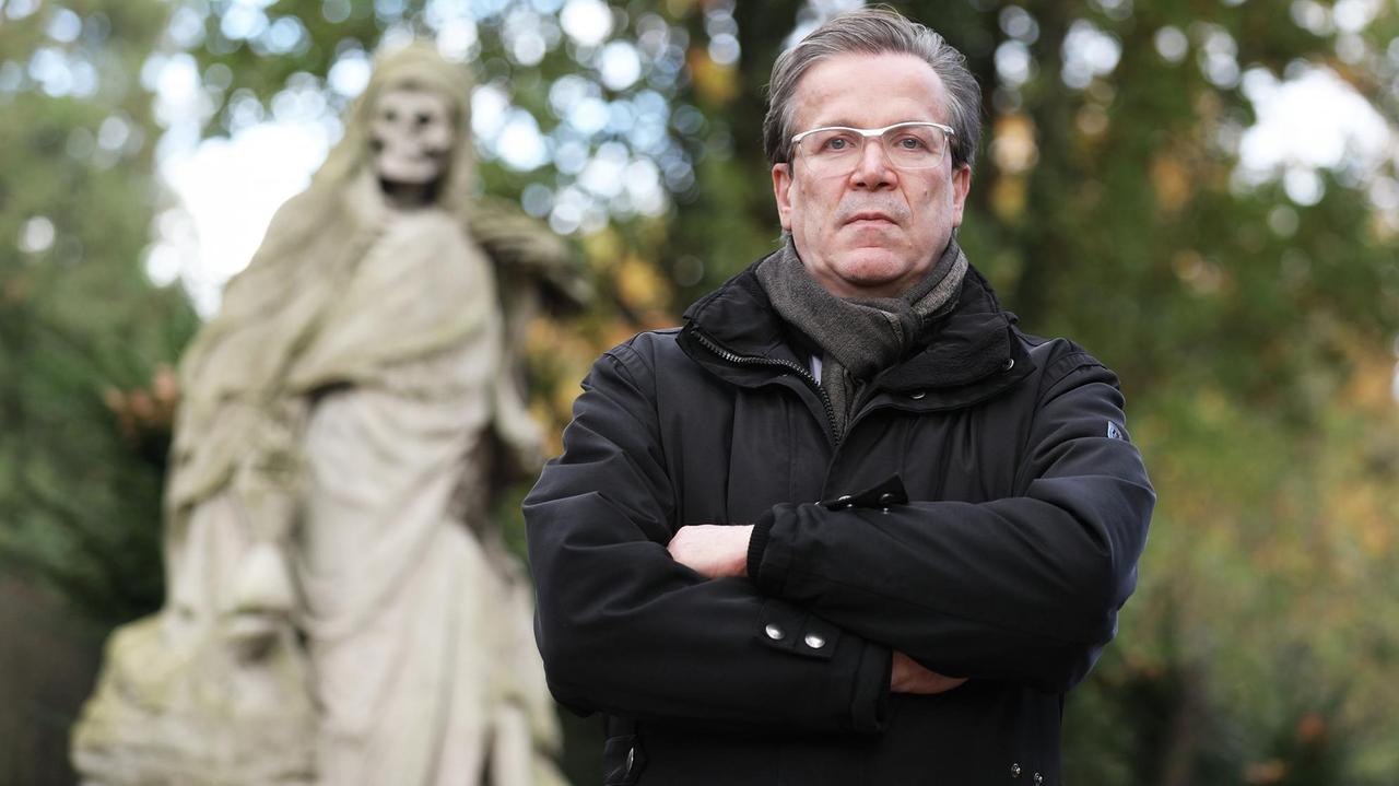 Christoph Kuckelkorn, Bestattungsunternehmer und Karnevalspräsident, steht auf einem Friedhof, links hinter ihm ein Grab mit einer Statue, die den Tod darstellt Christoph Kuckelkorn, Bestattungsunternehmer und Karnevalspräsident, steht auf einem Friedhof, links hinter ihm ein Grab mit einer Statue, die den Tod darstellt