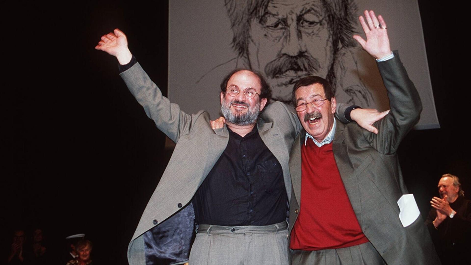 Salman Rushdie und Günter Grass stehen Arm in Arm auf einer Bühne und winken (1997)