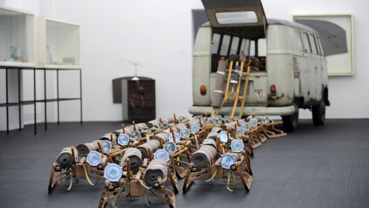 Ein Farbfoto zeigt die Installation "Das Rudel" von Joseph Beuys, bestehend aus 24 Schlitten, die vermeintlich aus der Heckklappe eines alten, gräulichen VW-Buses schwären. Auf jedem Schlitten befinden sich jeweils eine kleine Menge Fett, sowie eine Stablampe und eine eingerollte Filzdecke