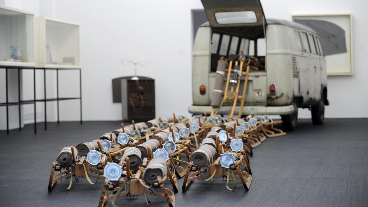 Ein Farbfoto zeigt die Installation "Das Rudel" von Joseph Beuys, bestehend aus 24 Schlitten, die vermeintlich aus der Heckklappe eines alten, gräulichen VW-Buses schwären. Auf jedem Schlitten befinden sich jeweils eine kleine Menge Fett, sowie eine Stablampe und eine eingerollte Filzdecke Ein Farbfoto zeigt die Installation "Das Rudel" von Joseph Beuys, bestehend aus 24 Schlitten, die vermeintlich aus der Heckklappe eines alten, gräulichen VW-Buses schwären. Auf jedem Schlitten befinden sich jeweils eine kleine Menge Fett, sowie eine Stablampe und eine eingerollte Filzdecke