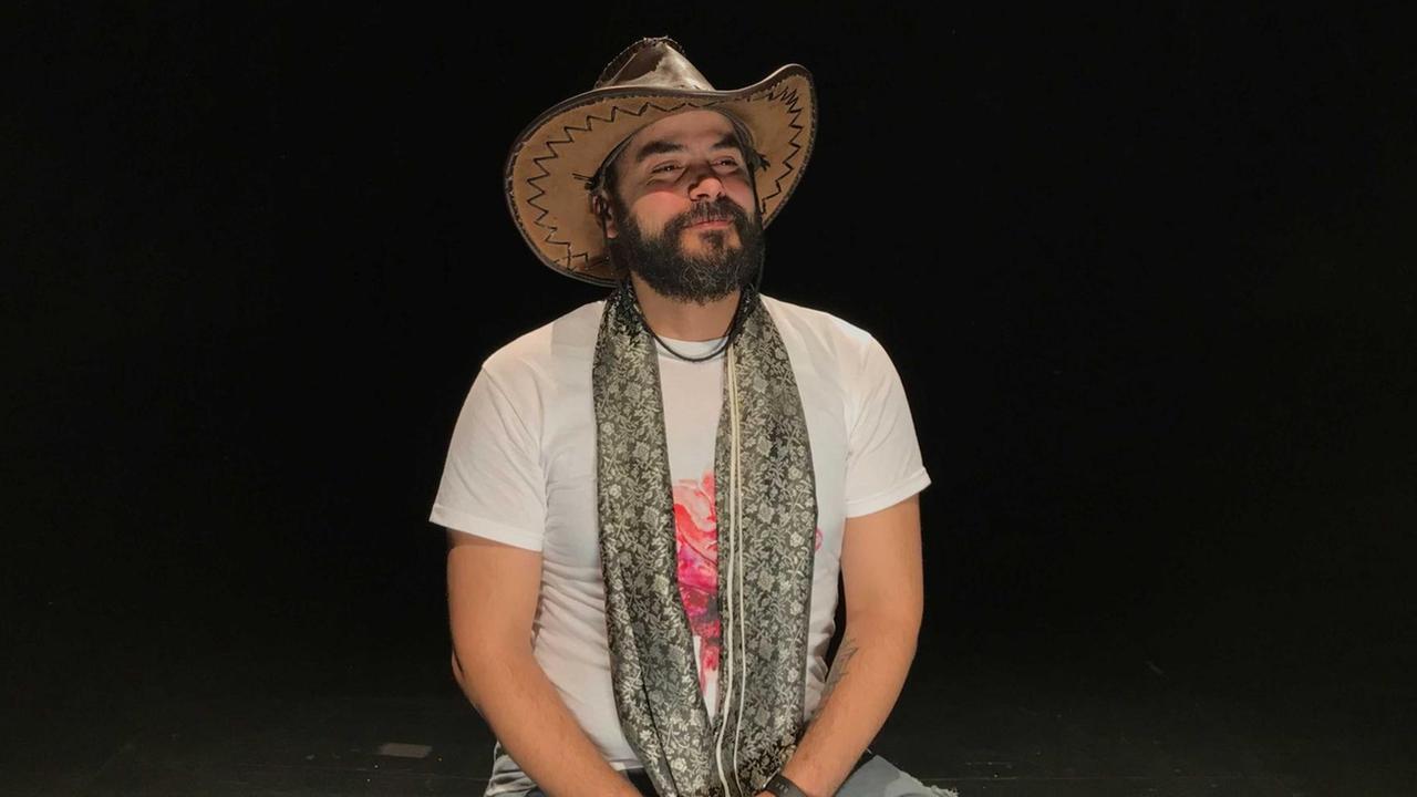 Der Kurator auf Kampnagel, Anas Aboura, auf einer dunklen Bühne sitzend, mit Jeans und weißem T-Shirt, einem Cowboy ähnlichen Hut und einem gemusterten Schal.