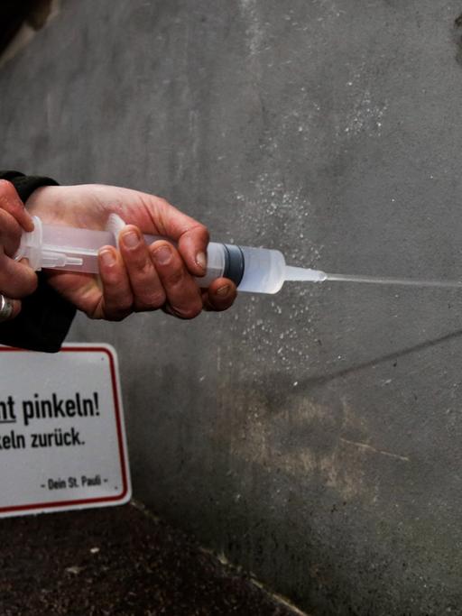 Die Quartiermanagerin der Reeperbahn, Julia Staron, demonstriert am 03.03.2015 in Hamburg im Stadtteil St. Pauli in der Einfahrt einer Tiefgarage neben einem Schild mit der Aufschrift "Hier nicht pinkeln! Wir pinkeln zurück" und einem Kanister mit superhydrophobem Lack die Reflexion von Flüssigkeit. Weil Betrunkene in dem legendären Stadtteil überall hinpinkeln, hat die Interessengemeinschaft St. Pauli Wände, an die in dem Viertel oft gepinkelt wird, mit superhydrophobem Lack beschichtet, der so wasserabweisend ist, dass der Urin regelrecht abprallt und zurückgespritzt wird.