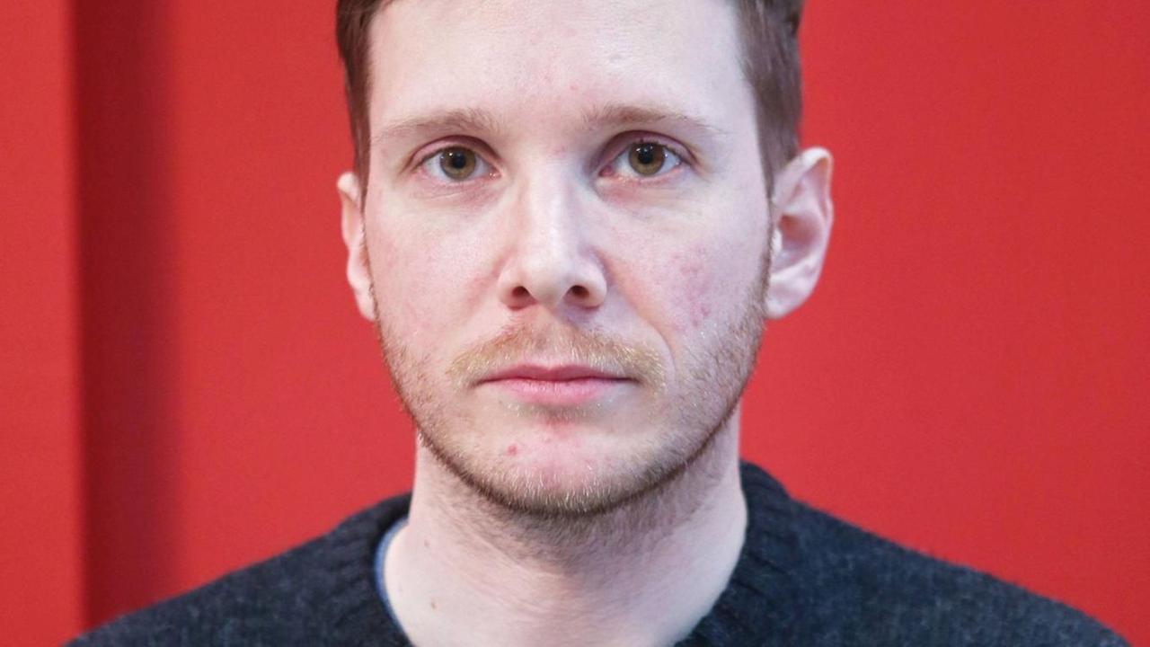 Porträtfoto eines junge Mannes mit grauem Strickpulli vor einer roten Wand. Der Mann schaut moderat-traurig in die Kamera, mit großen braunen Augen. Porträtfoto eines junge Mannes mit grauem Strickpulli vor einer roten Wand. Der Mann schaut moderat-traurig in die Kamera, mit großen braunen Augen.