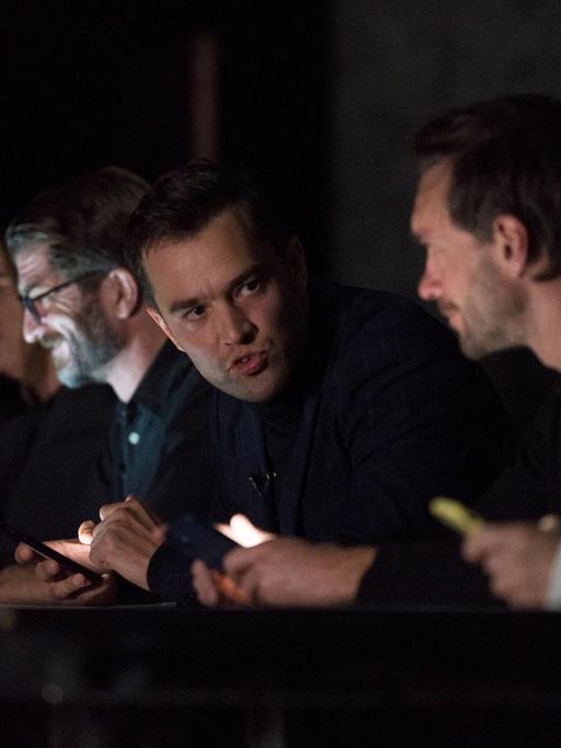 Sechs Schauspieler sitzen an einem langen Tisch und starren in ihre Handys. Nils Strunk, der Thomas Schmid spielt, ist hell erleuchtet.