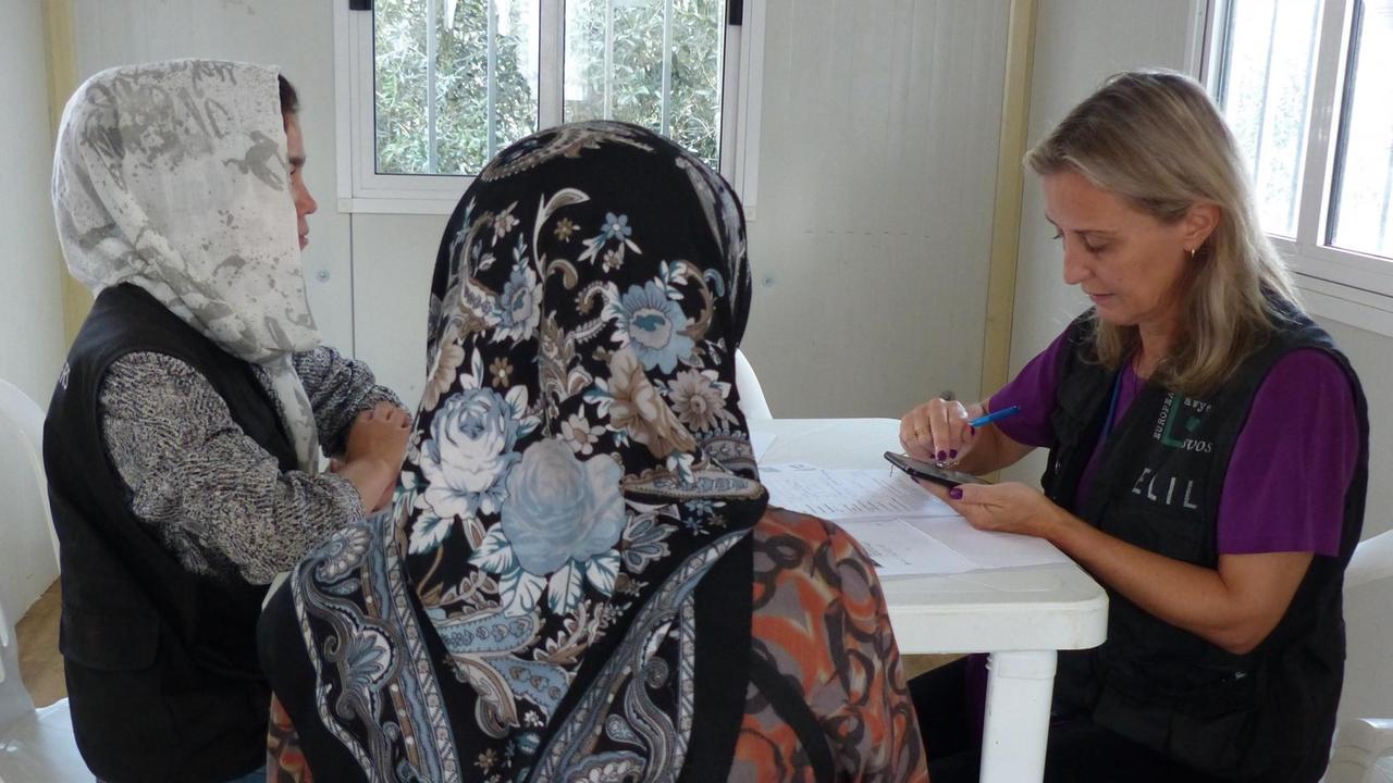 Zwei Frauen mit Kopftuch sitzen an einem weißen Tisch einer jungen Frau gegenüber, die in ihr Smartphone schaut. Zwei Frauen mit Kopftuch sitzen an einem weißen Tisch einer jungen Frau gegenüber, die in ihr Smartphone schaut.