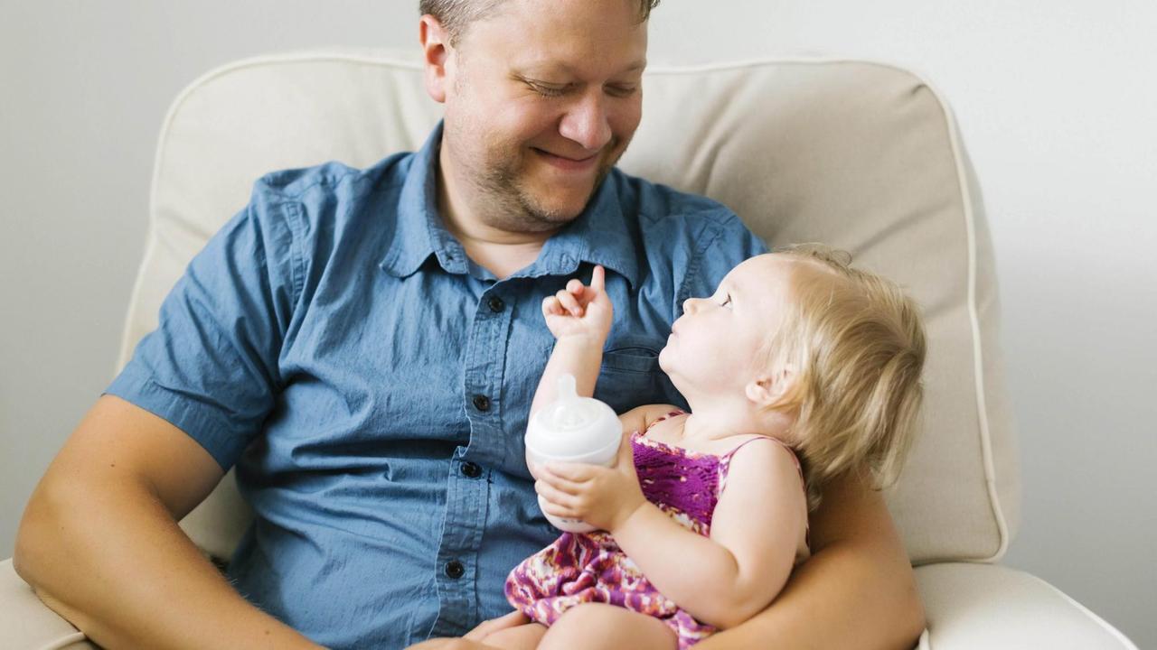 Ein Vater mit einem Baby mit Trinkflasche auf dem Schoß, er lächelt sie an, die Kleine zeigt auf ihn.