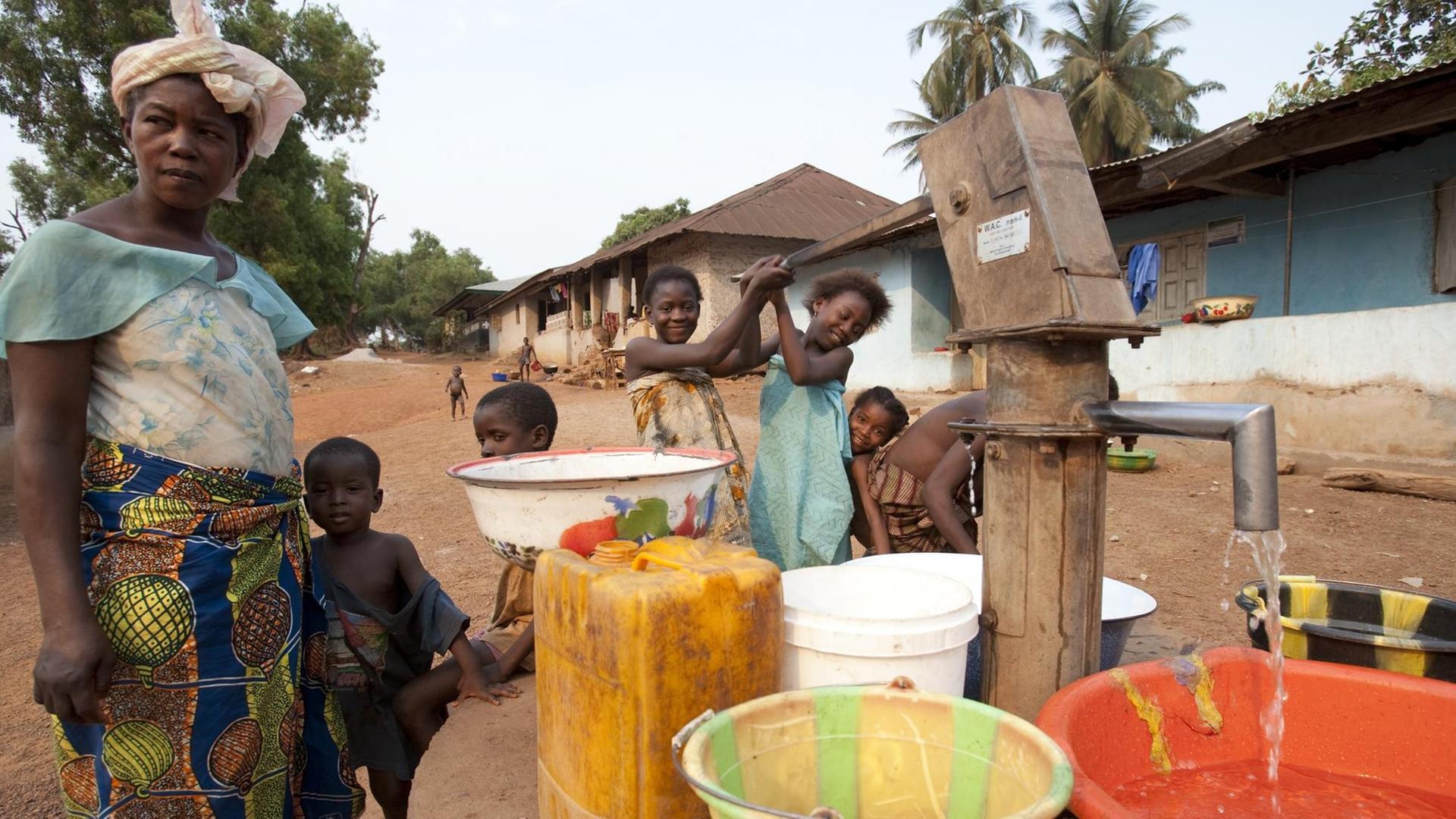 Szene aus der Stadt Makeni, im Nordosten von Sierra Leone: Kinder pumpen sauberes Wasser aus einem Brunnen in Schüsseln und Kanister. Links daneben steht eine Frau in einem bunten Rock. Szene aus der Stadt Makeni, im Nordosten von Sierra Leone: Kinder pumpen sauberes Wasser aus einem Brunnen in Schüsseln und Kanister. Links daneben steht eine Frau in einem bunten Rock.