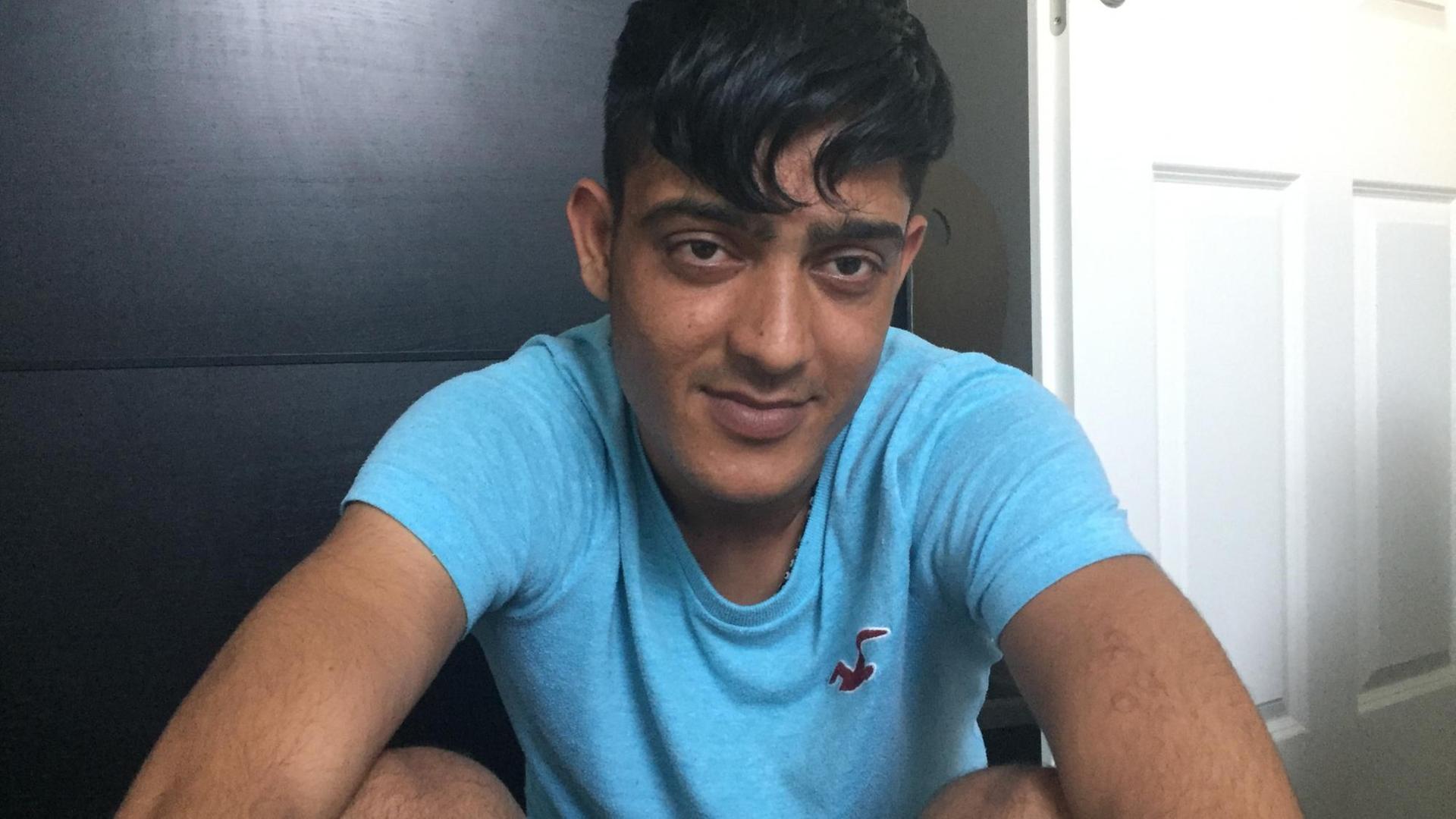 Der afghanische Flüchtling Attaullah Baloz sitzt auf dem Boden, hinter ihm ist eine weiße Holztür und eine schwarze Holzwand zu sehen. Er trägt ein türkisfarbenes T-Shirt Der afghanische Flüchtling Attaullah Baloz sitzt auf dem Boden, hinter ihm ist eine weiße Holztür und eine schwarze Holzwand zu sehen. Er trägt ein türkisfarbenes T-Shirt