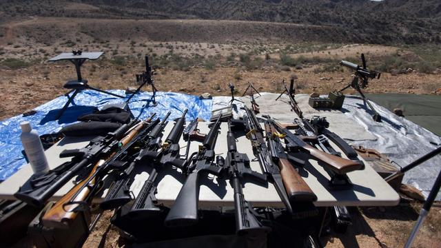 Auf dem Tisch in einer Wüstenlandschaft liegen zahlreiche Gewehre nebeneinander.