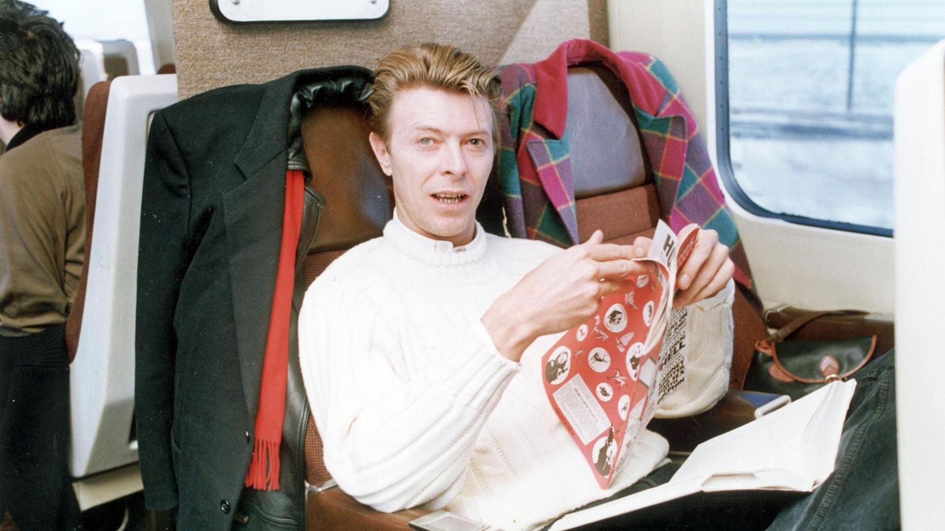 David Bowie sitzt im Zug, auf seinem Schoß ein Buch, in den Händen hält er eine Zeitschrift. Er schaut in die Kamera. David Bowie sitzt im Zug, auf seinem Schoß ein Buch, in den Händen hält er eine Zeitschrift. Er schaut in die Kamera.