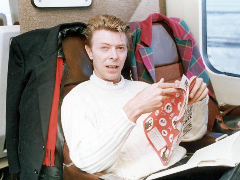 David Bowie sitzt im Zug, auf seinem Schoß ein Buch, in den Händen hält er eine Zeitschrift. Er schaut in die Kamera.