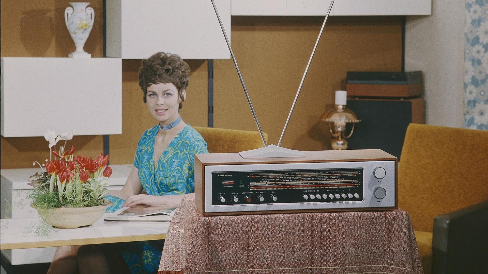 Eine junge Frau sitzt in einem Siebzigerjahre-Wohnzimmer an einem Tisch, im Vordergrund steht ein Radiogeraet REMA adagio.