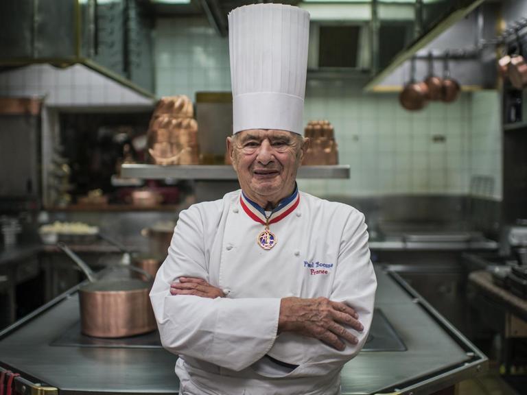 Paul Bocuse steht im Jahr 2012 mit verschränkten Armen in der Küche und lächelt in die Kamera.