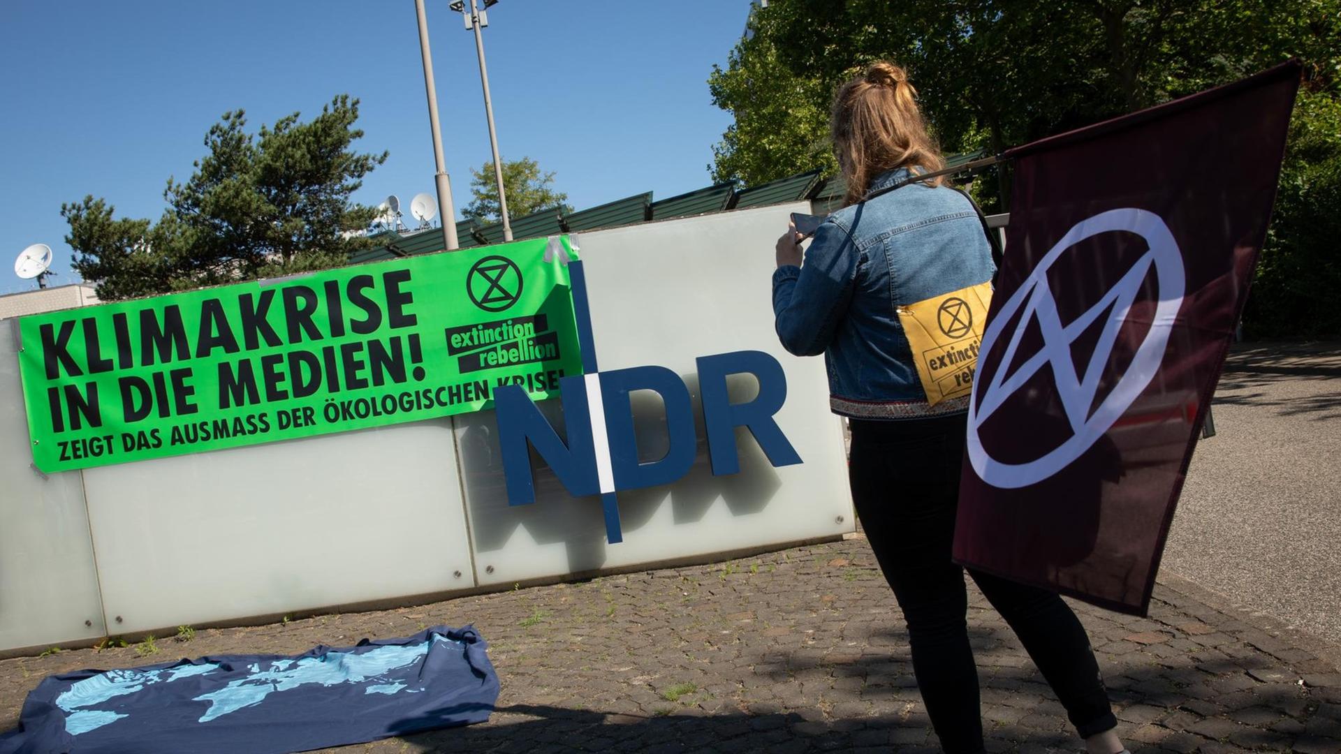 5.06.2020, Hamburg: Eine Aktivistin der Umweltschutzbewegung Extinction Rebellion (XR) steht während eines Protests mit einer Flagge vor dem Eingang zum Norddeutschen Rundfunk (NDR), an dessen Mauer ein Banner mit der Aufschrift «Klimakrise in die Medien! Zeigt das Ausmaß der ökologischen Krise» hängt. Mit einer Blockade vor der Einfahrt zum NDR-Gelände in Hamburg-Lokstedt haben die Umweltaktivisten den Sender aufgefordert, in seiner Berichterstattung den Begriff Klimawandel durch Klimakrise zu ersetzen. Foto: Christian Charisius/dpa 5.06.2020, Hamburg: Eine Aktivistin der Umweltschutzbewegung Extinction Rebellion (XR) steht während eines Protests mit einer Flagge vor dem Eingang zum Norddeutschen Rundfunk (NDR), an dessen Mauer ein Banner mit der Aufschrift «Klimakrise in die Medien! Zeigt das Ausmaß der ökologischen Krise» hängt. Mit einer Blockade vor der Einfahrt zum NDR-Gelände in Hamburg-Lokstedt haben die Umweltaktivisten den Sender aufgefordert, in seiner Berichterstattung den Begriff Klimawandel durch Klimakrise zu ersetzen. Foto: Christian Charisius/dpa