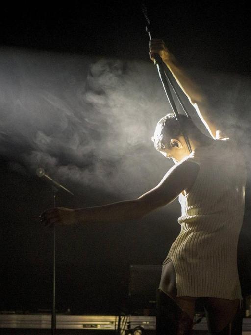Arca bei einem Auftritt auf der Bühne mit dem Rücken zur Kamera, in einem Lichtstrahl mit Nebel