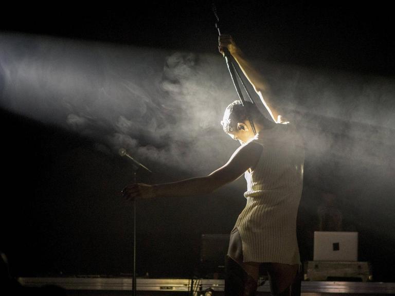 Arca bei einem Auftritt auf der Bühne mit dem Rücken zur Kamera, in einem Lichtstrahl mit Nebel