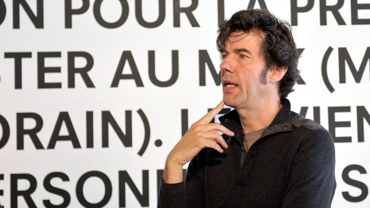 Stefan Sagmeister, schwarzes Hemd, schwarze Haare, steht redend vor einer weißen Wand mit schwarzen, französischen Worten.