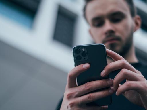 Von unten fotografierter junger Mann, der in seinen Händen ein Smartphone hält und auf dessen Bildschirm blickt.