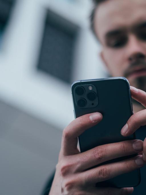 Von unten fotografierter junger Mann, der in seinen Händen ein Smartphone hält und auf dessen Bildschirm blickt.