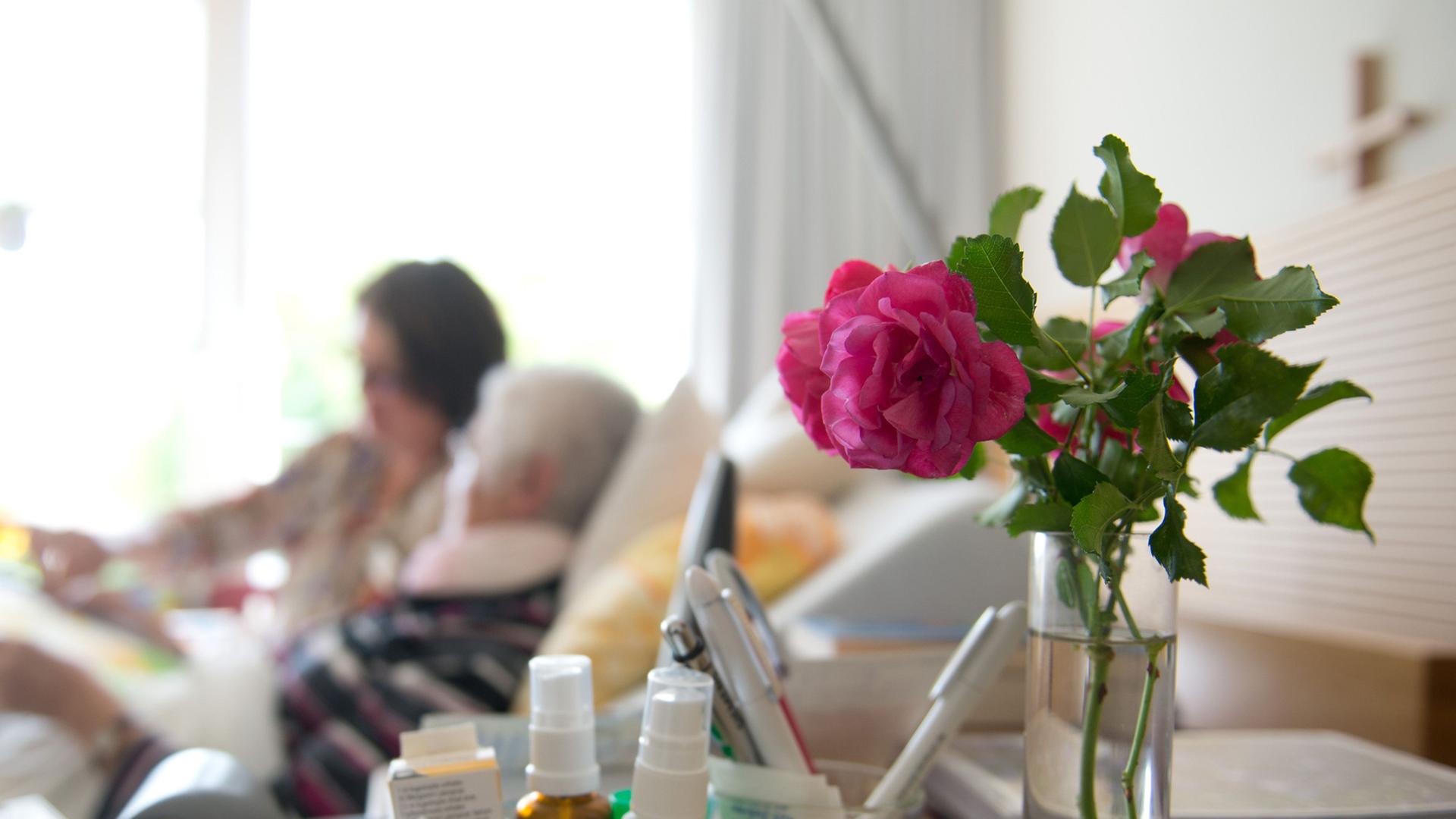 Im Vordergrund eine Rose, im Hintergrund ein Krankenbett mit einer alten Frau und einer jüngeren am Bett. Im Vordergrund eine Rose, im Hintergrund ein Krankenbett mit einer alten Frau und einer jüngeren am Bett.
