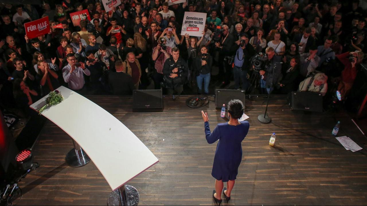 Sahra Wagenknecht steht auf einer Bühne und spricht zur Besuchern der Wahlparty der Linken. In der Menge halten Menschen Plakate der Linken hoch. Sicht von oben.