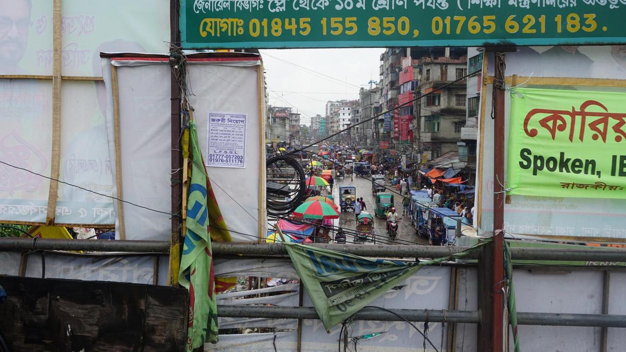 Durch ein Loch in der Rückseite eines Plakats schauen wir auf eine viel befahrene Straße in der Innenstadt von Dhaka.
