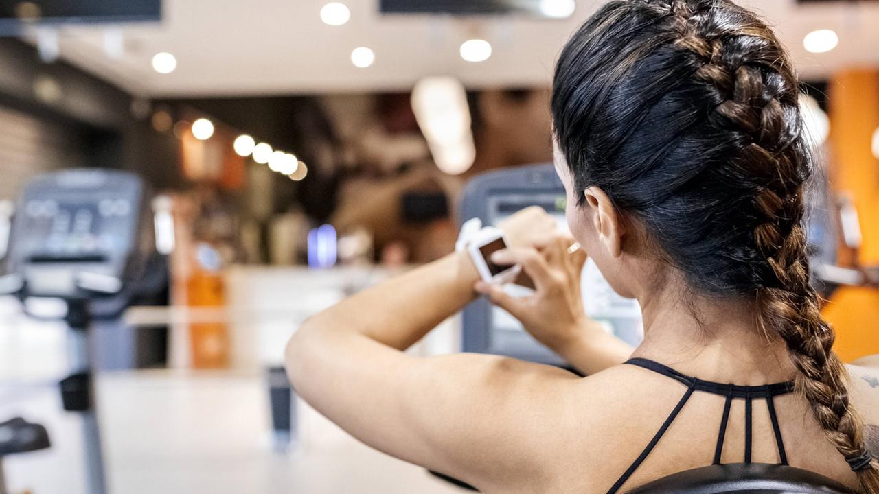 Rückansicht eine Frau im Fitnesstudio, die auf ihre Smartwatch schaut. Auf der Schulter rechts hat sie mehrere Tätowierungen.