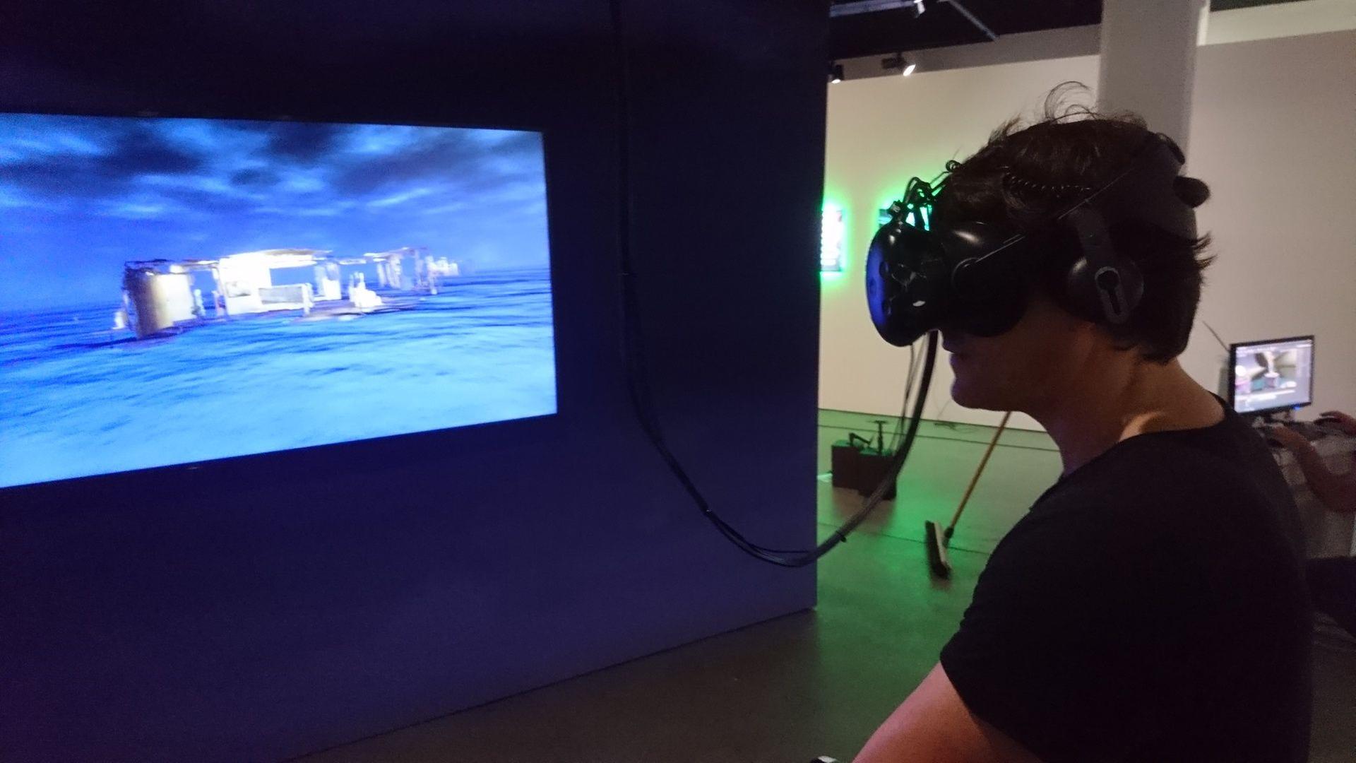 Ein Mann sitzt in einer relativ dunklen Ausstellungshalle, er ist in Umrissen zu erkennen. Vor ihm ist ein Bild aus einem Game zu sehen, das er gerade spielt Ein Mann sitzt in einer relativ dunklen Ausstellungshalle, er ist in Umrissen zu erkennen. Vor ihm ist ein Bild aus einem Game zu sehen, das er gerade spielt