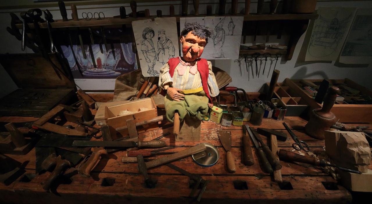 Eine Marionettenfigur auf einem Schreinertisch mit Werkzeugen drumherum, im Hintergrund sind Zeichnung zu sehen.