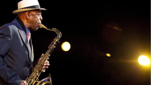 Der Saxofonist Archie Shepp steht auf einer Bühne und spielt sein Instrument, auf der rechten Seite ist ein Scheinwerfer zu sehen, der den Musiker anstrahlt.
