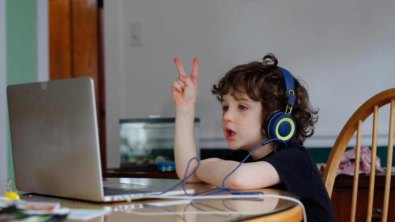 Ein Junge mit Kopfhörern sitzt vor einem Laptop, er streckt fröhlich eine Hand mit dem Victoryzeichen in die Luft. Ein Junge mit Kopfhörern sitzt vor einem Laptop, er streckt fröhlich eine Hand mit dem Victoryzeichen in die Luft.