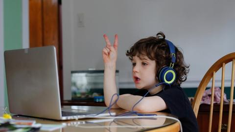 Ein Junge mit Kopfhörern sitzt vor einem Laptop, er streckt fröhlich eine Hand mit dem Victoryzeichen in die Luft.