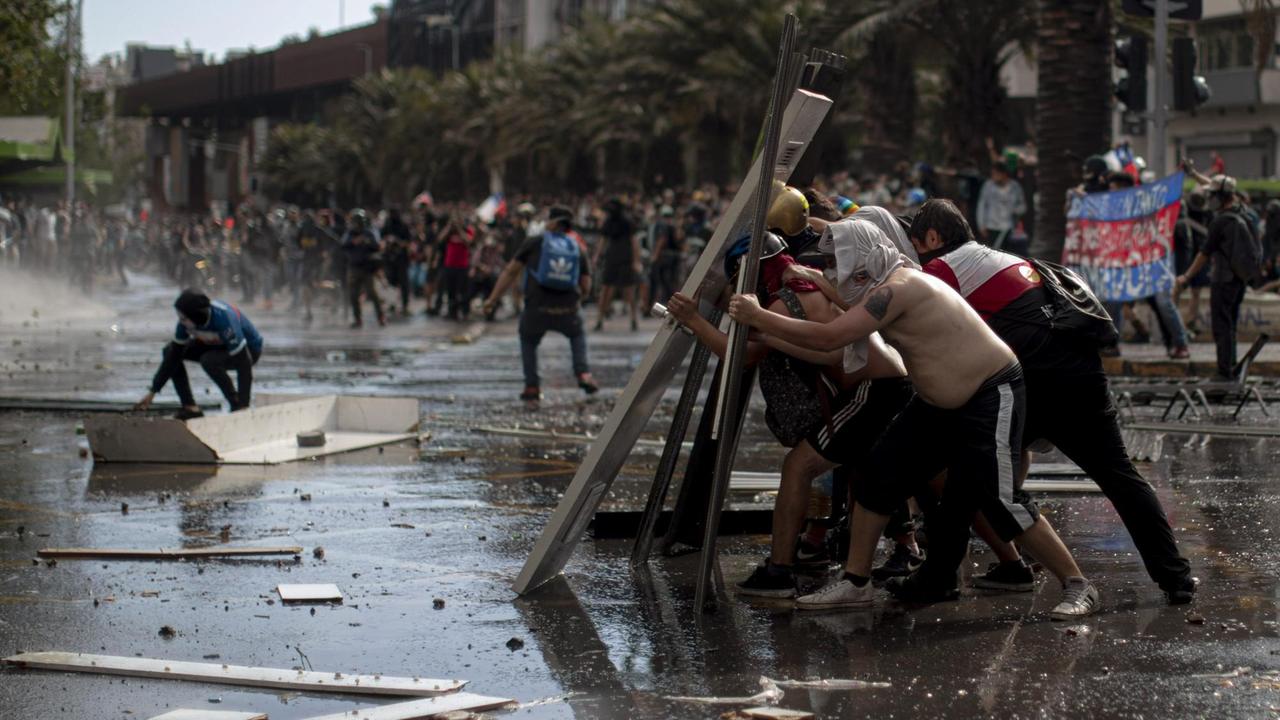 Szenen einer gewaltvollen Demonstration in den Straßen von Santiago de Chile. Im Vordergrund Demonstranten, die sich mit Türen schützen, im Hintergrund eine Masse von Menschen im Tumult.
