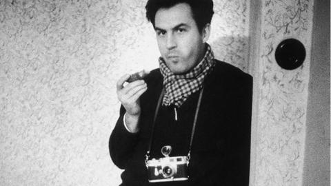 Ein Schwarzweiß-Foto zeigt den österreichischen Fotografen Ernst Hans mit umgehängter Kamera in schwarzem Pullover und mit kariertem Schal an einen Türrahmen gelehnt.