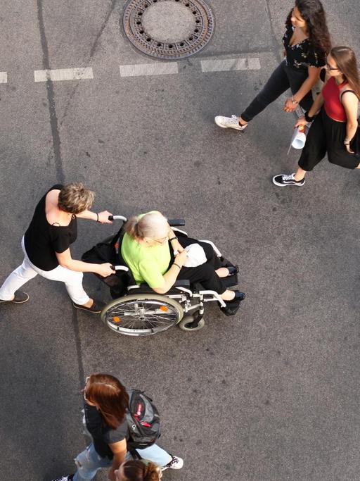 Das Bild zeigt eine Straße von oben mit Menschen, in der Mitte eine Frau im Rollstuhl.