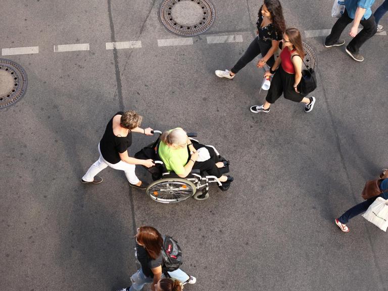 Das Bild zeigt eine Straße von oben mit Menschen, in der Mitte eine Frau im Rollstuhl.