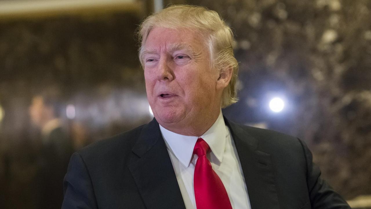Trump steht vor einer dunklen Wand und deutet mit dem Zeigerfinger nach vorne. Trump steht vor einer dunklen Wand und deutet mit dem Zeigerfinger nach vorne.