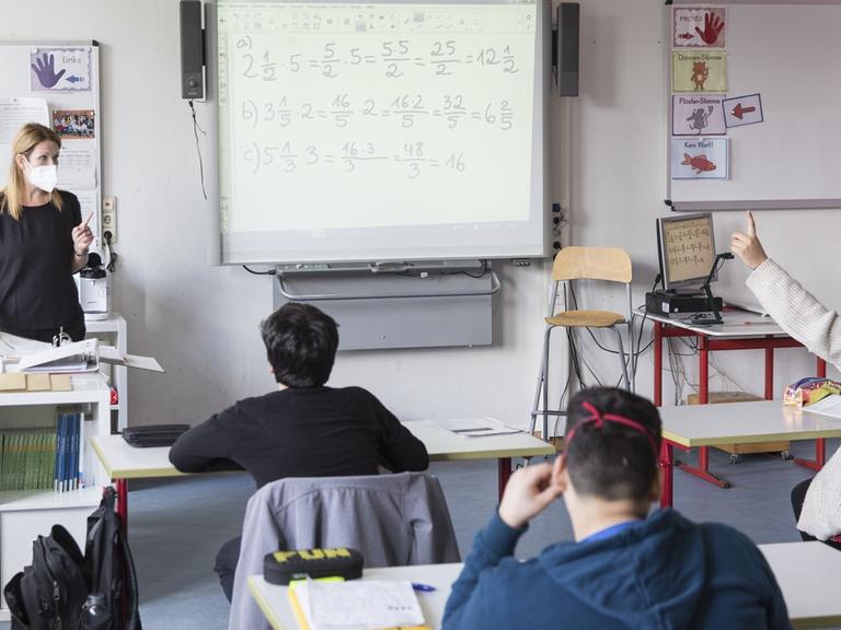 Schüler sitzen vor ihrer Lehrerin im Klassenraum, neben der Lehrerin steht ein Whiteboard, daneben ein Computer.