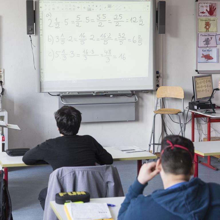 Schüler sitzen vor ihrer Lehrerin im Klassenraum, neben der Lehrerin steht ein Whiteboard, daneben ein Computer.