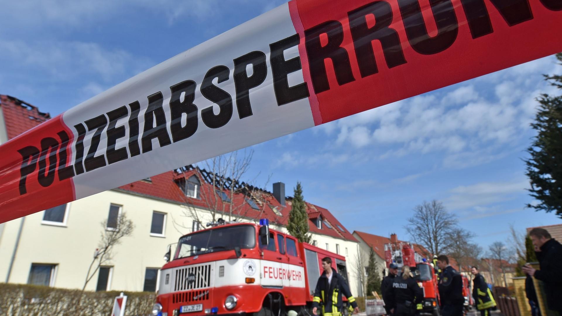 Ein weiß-rotes Band mit der Aufschrift "Polizeiabsperrung" hängt quer über die Straße. Dahinter sieht man ein Feuerwehrauto mit Feuerwehrmännern und Polizisten sowie das Heim mit dem abgebrannten Dach. Ein weiß-rotes Band mit der Aufschrift "Polizeiabsperrung" hängt quer über die Straße. Dahinter sieht man ein Feuerwehrauto mit Feuerwehrmännern und Polizisten sowie das Heim mit dem abgebrannten Dach.