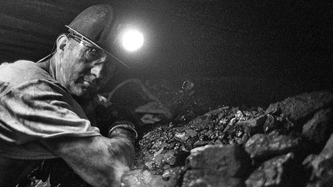 Ein Bergmann arbeitet auf der Zeche Prosper- Haniel an einem Flöz unter Tage vor Kohle. Auf seinem Helm eluchtet eine Lampe. Das Bild ist schwarz-weiß.