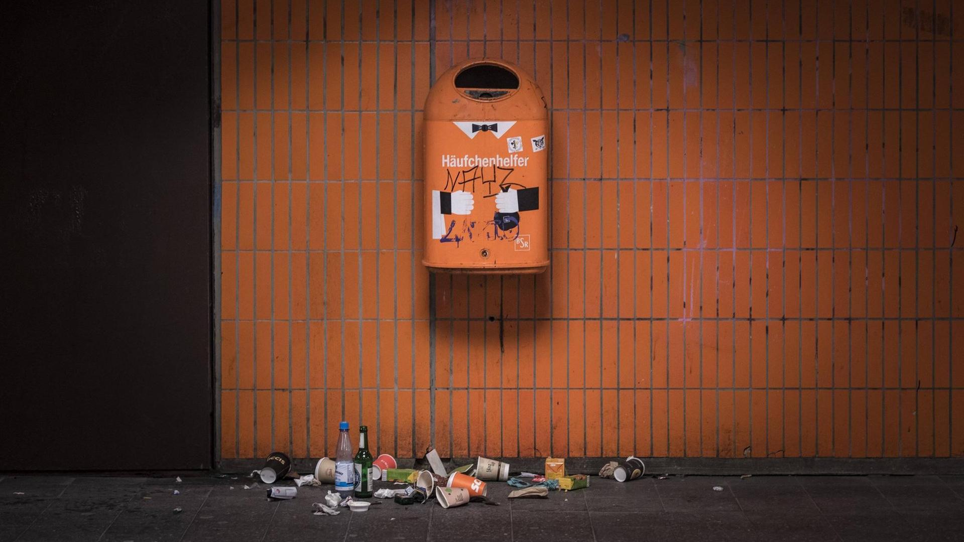 Ein orangener Mülleimer an einer orang gekachelten Wand in Berlin. Darauf steht Häufchenhelfer. Darunter liegt verschiedenartiger Abfall. Ein orangener Mülleimer an einer orang gekachelten Wand in Berlin. Darauf steht Häufchenhelfer. Darunter liegt verschiedenartiger Abfall.