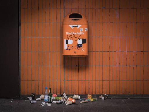 Ein orangener Mülleimer an einer orang gekachelten Wand in Berlin. Darauf steht Häufchenhelfer. Darunter liegt verschiedenartiger Abfall.