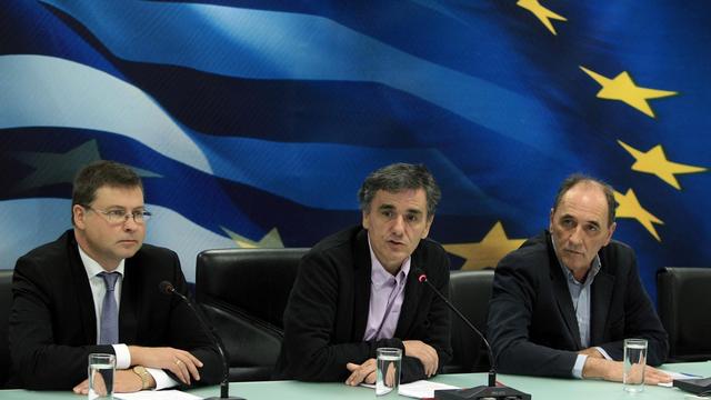 Dombrovskis, Tsakalotos und Stathakis sitzen an einem Pult. Tsakalatos spricht in ein Mikrofon. Die drei sitzen vor einer Wand, auf der die Fahnen Griechenlands und der EU zu sehen sind.